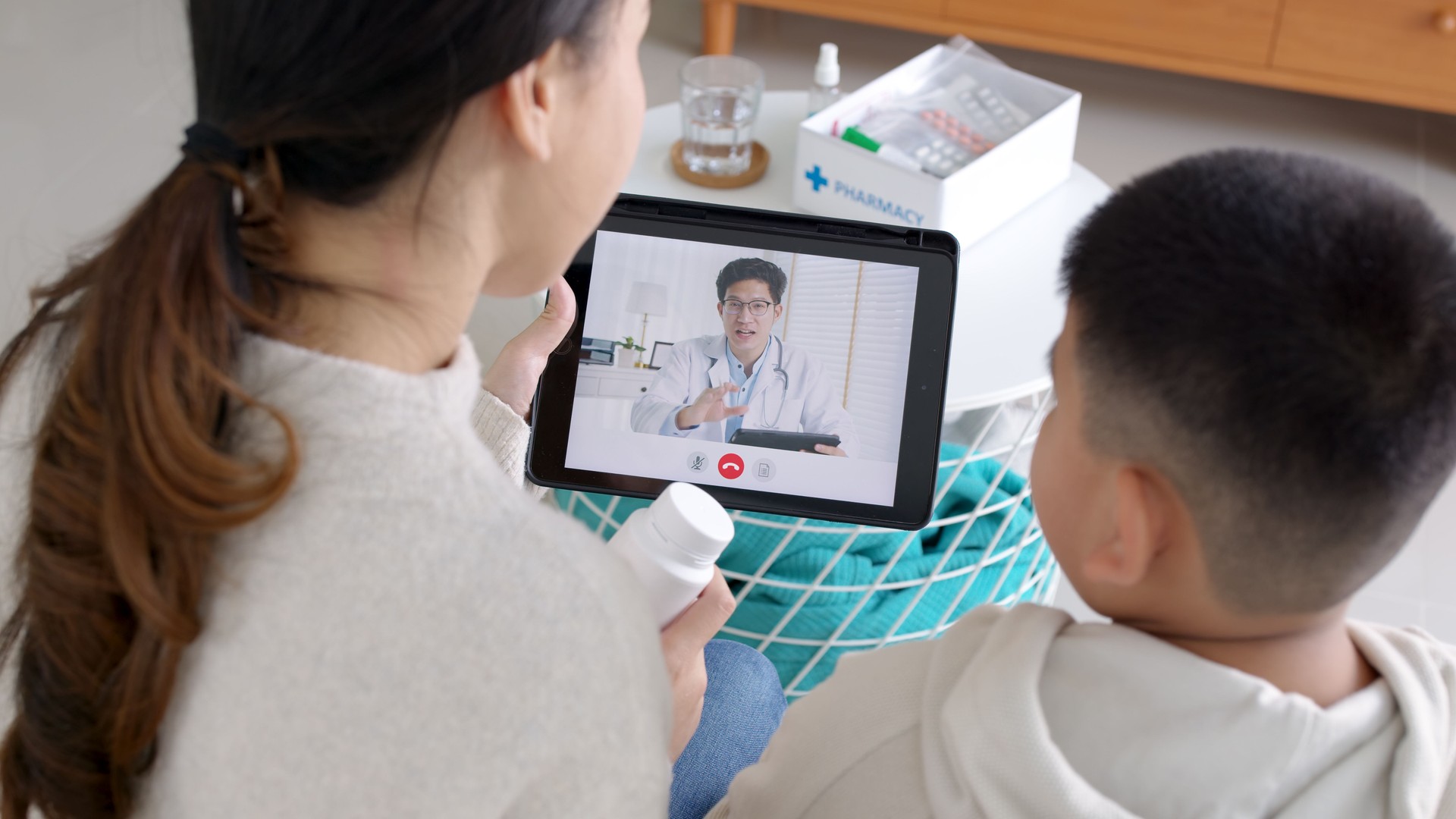 Over shoulder view of young asia parents and son talk to doctor on cellphone videocall conference medical app in telehealth telemedicine online service hospital quarantine social distance at home.
